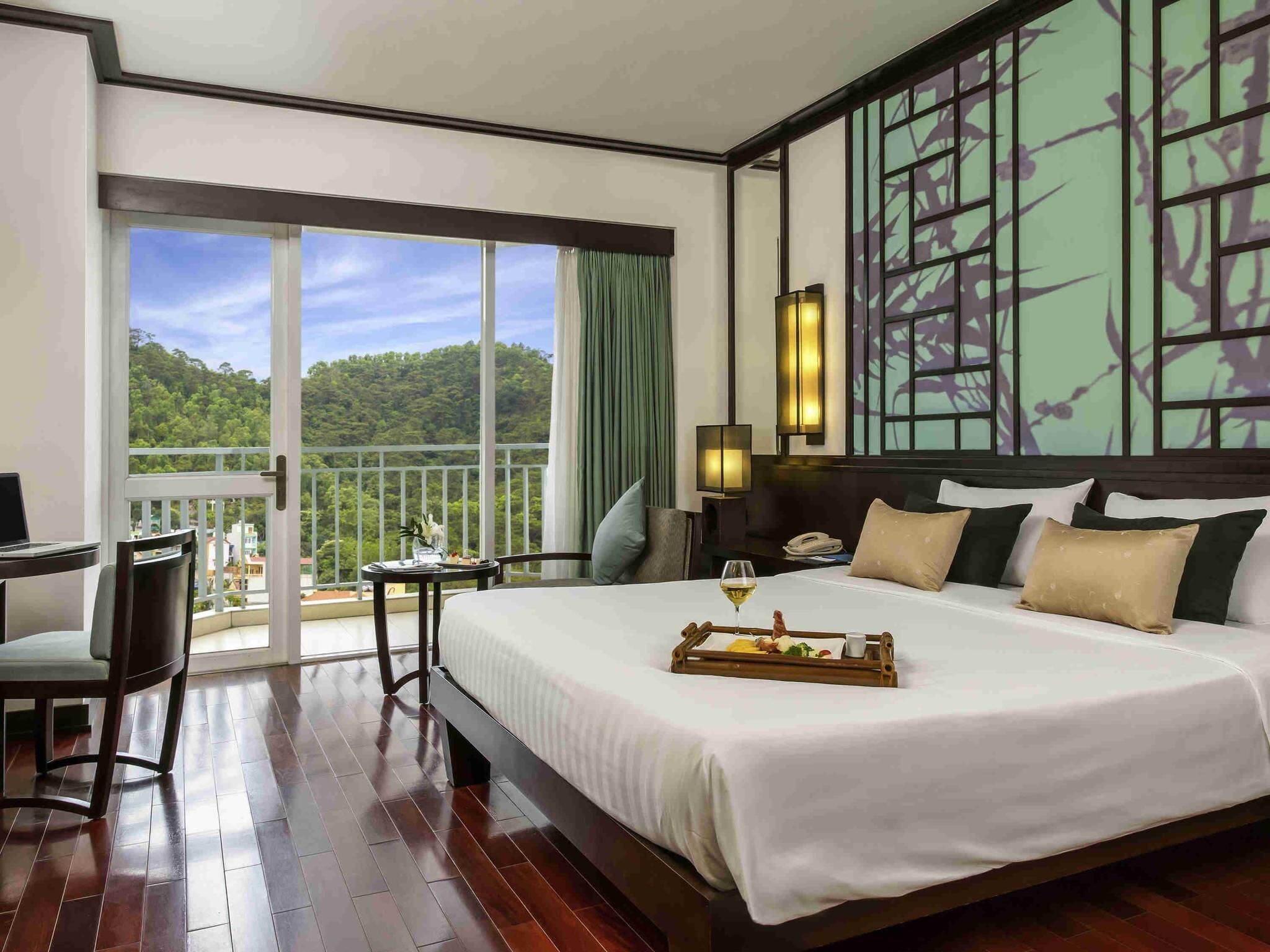 Habitación de hotel con una cama grande, un escritorio, una silla, un balcón con vistas al bosque y una pared con un panel decorativo.