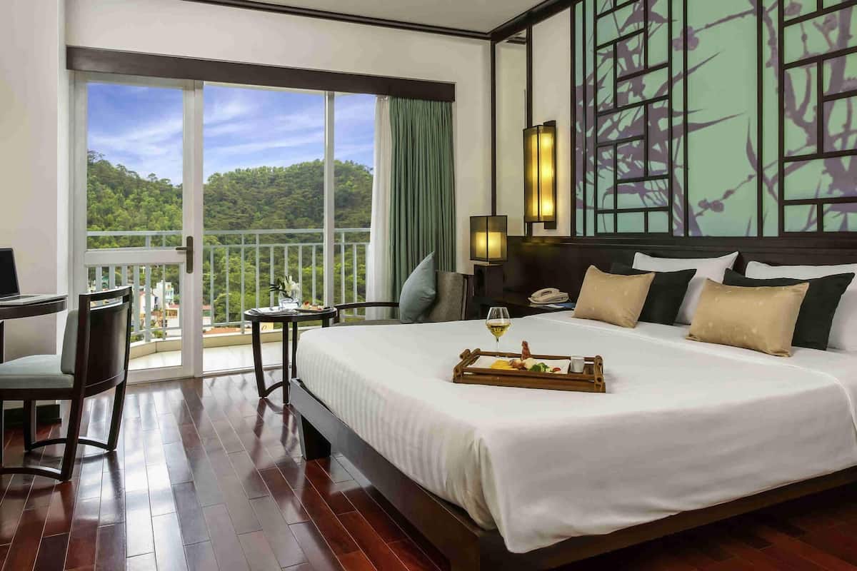 Habitación de hotel con una cama grande, un escritorio, una silla, un balcón con vistas al bosque y una pared con un panel decorativo.