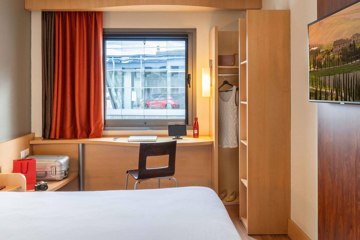 Une chambre d’hôtel de taille réduite, comprenant un lit, un bureau avec une chaise, une fenêtre avec des rideaux, un porte-manteau et un tableau accroché au mur.