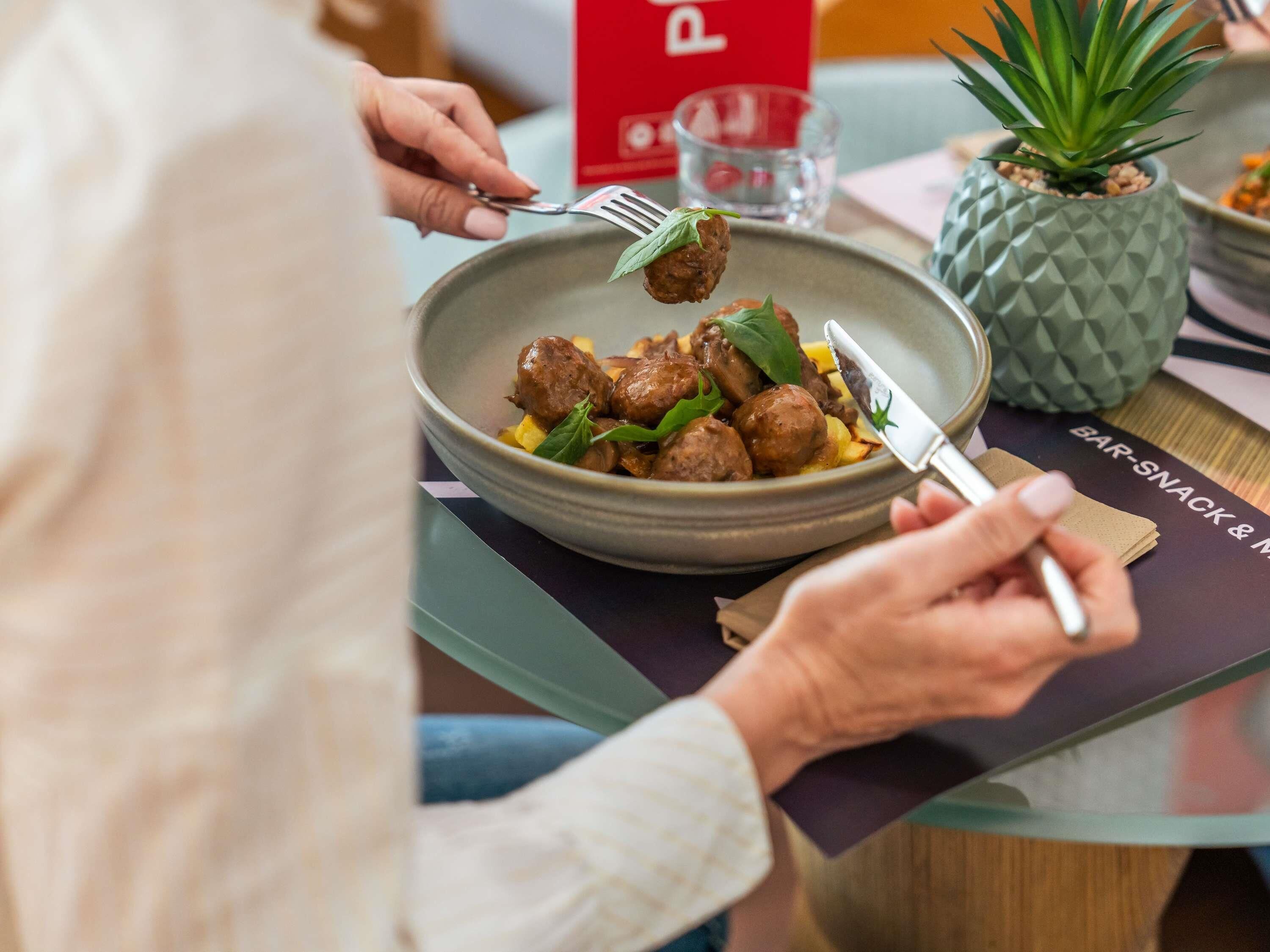 A person holding a fork with meatballs and greens on a plate, a knife, and a pineapple plant on a table.