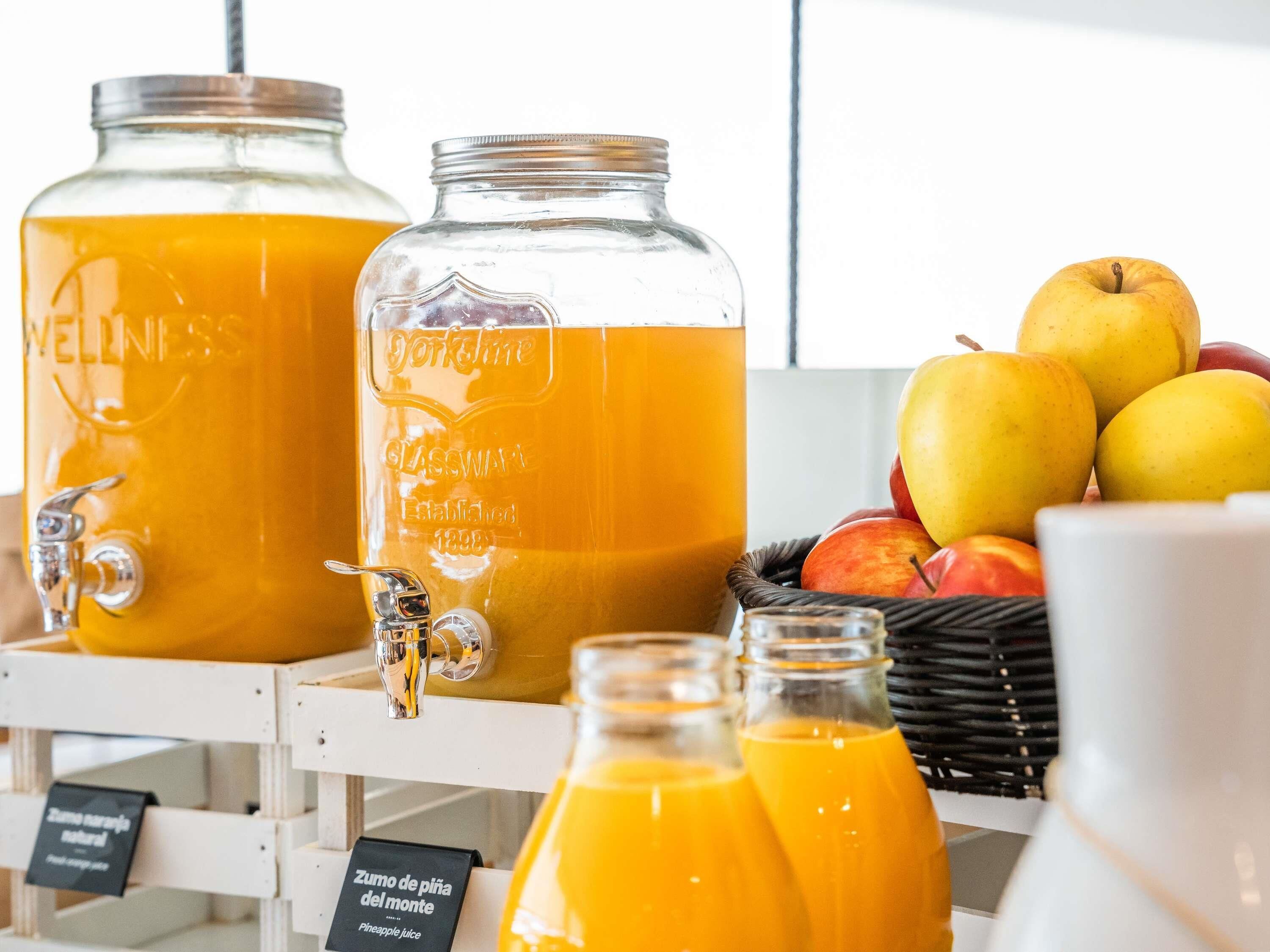 Dos grandes dispensadores de jugo de naranja, una cesta de manzanas y un cartel que indica 'Pineapple juice'.