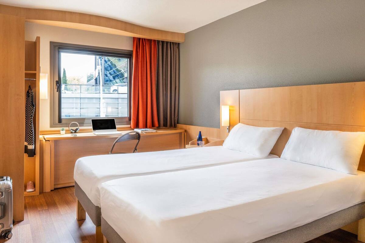 Une chambre d’hôtel avec deux lits, un bureau avec un ordinateur portable, une chaise, une fenêtre avec des rideaux et une valise posée sur le sol.