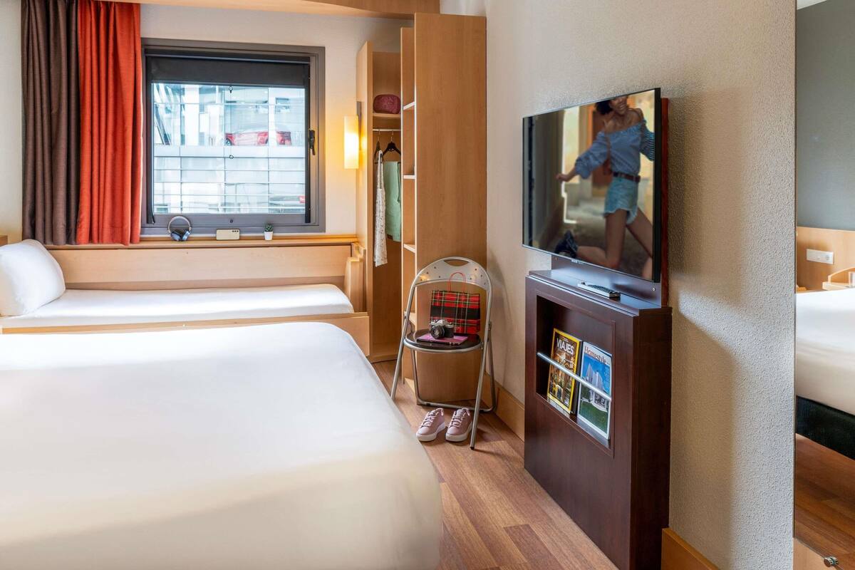 Une chambre d’hôtel avec deux lits, une télévision, une armoire et une fenêtre avec des rideaux.