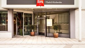 Entrada de hotel con puertas de vidrio, plantas en macetas y un letrero que indica 'ibis Madrid Alcobendas'.