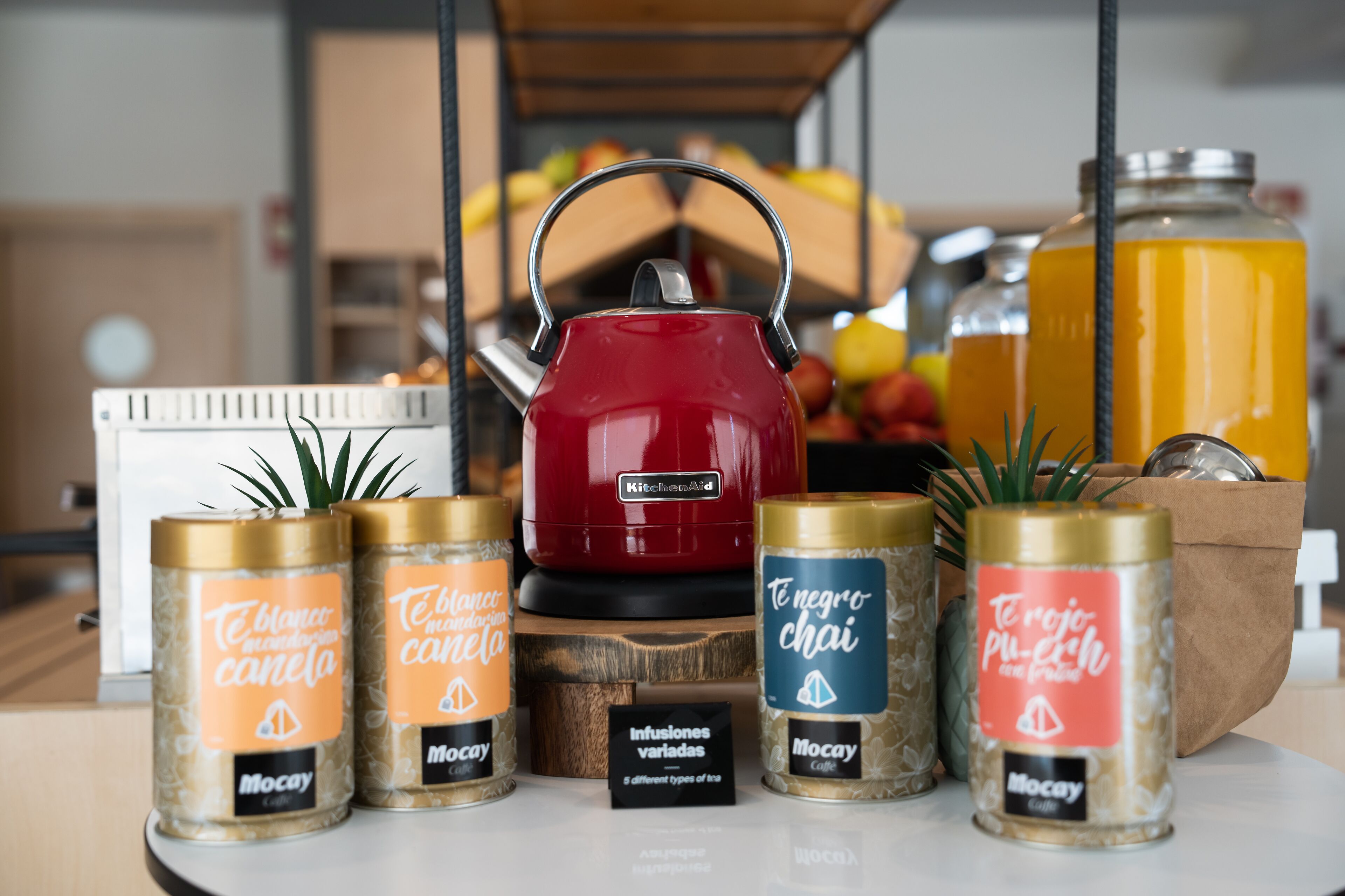 A kitchen counter with various items including a red kettle, jars of tea, and a large jar of orange juice.