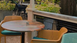 An outdoor patio with a round table and two wicker chairs, surrounded by greenery and a tree. - Hotel Bentheimer Hof (Bad Bentheim)