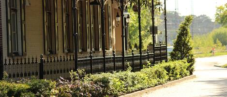 A building with a yellow facade, black iron fence, and a garden with various plants.