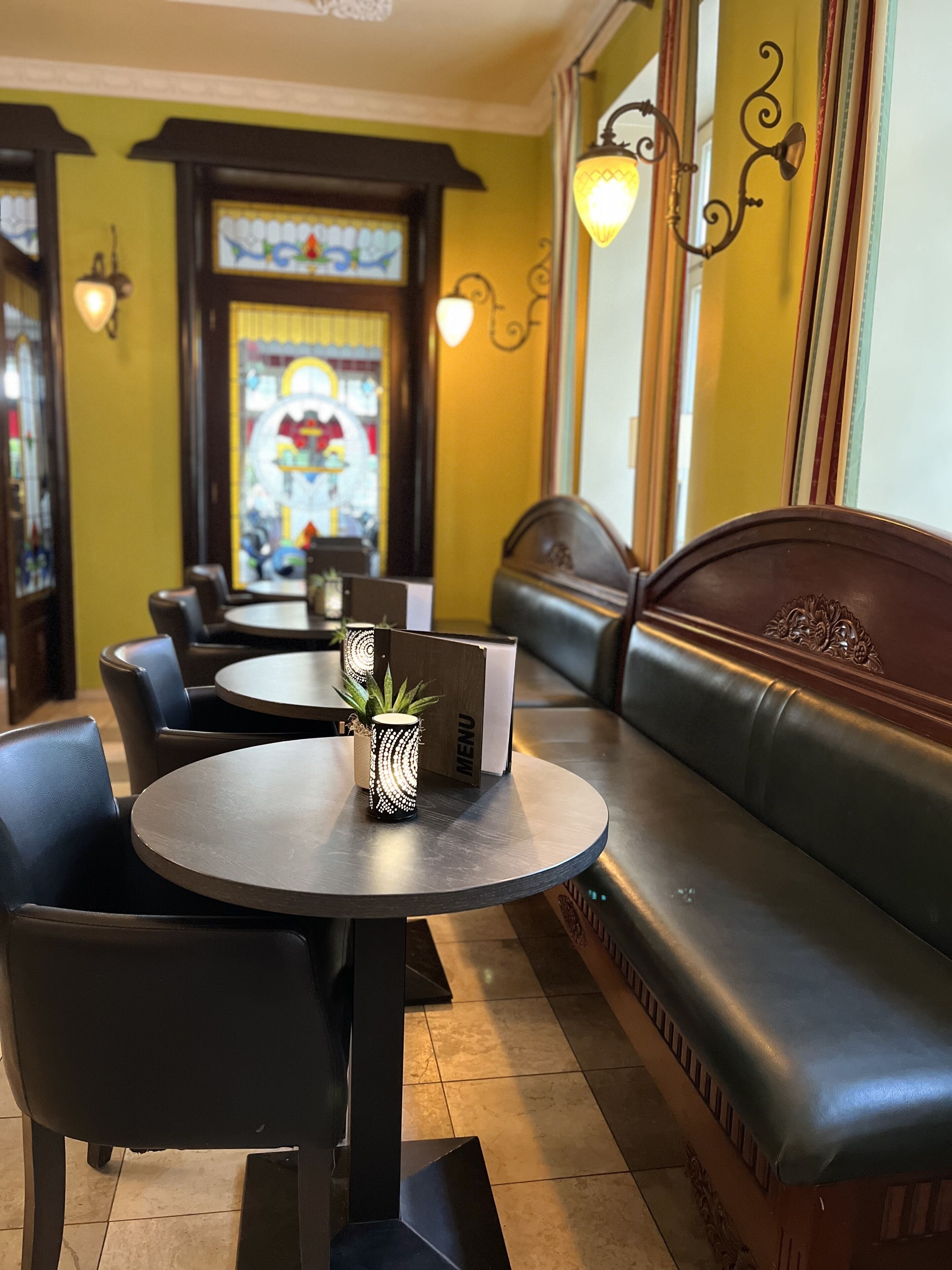 Ein Restaurantinnenraum mit runden Tischen, schwarzen Ledersitzgruppen und einem Buntglasfenster im Hintergrund.