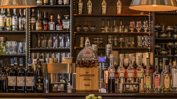 A bar area with various bottles of alcohol, a wooden bar counter, and shelves stocked with glasses and bottles in the background. - Hotel Bentheimer Hof (Bad Bentheim)