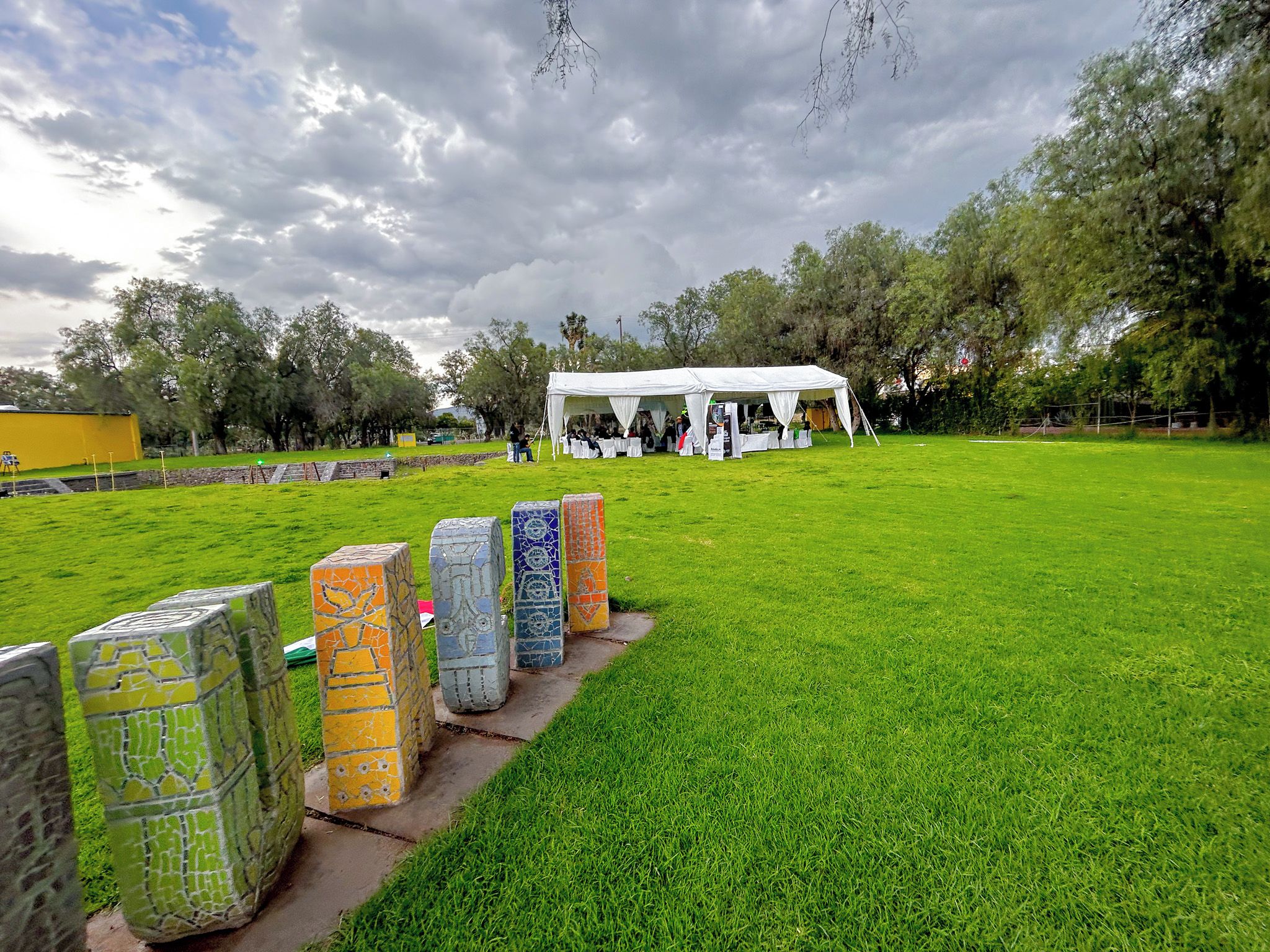 A grassy field with a white tent and a group of people. There are colorful mosaic barriers in the foreground.