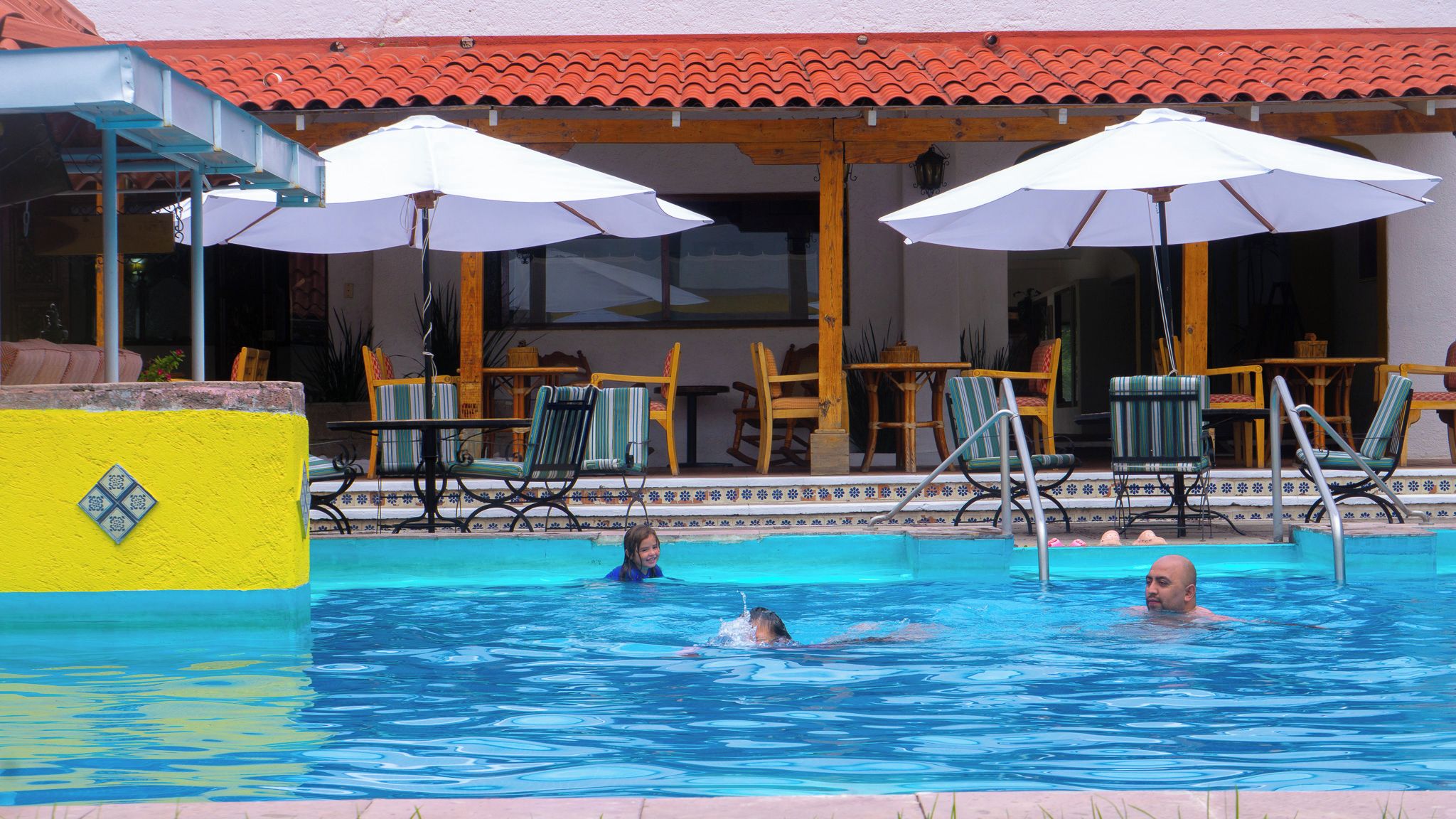 Una piscina con sillones de descanso y sombrillas, techo de baldosas y pared amarilla con detalles en cerámica.