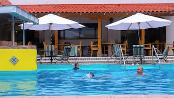 Una piscina con sillones de descanso y sombrillas, techo de baldosas y pared amarilla con detalles en cerámica.