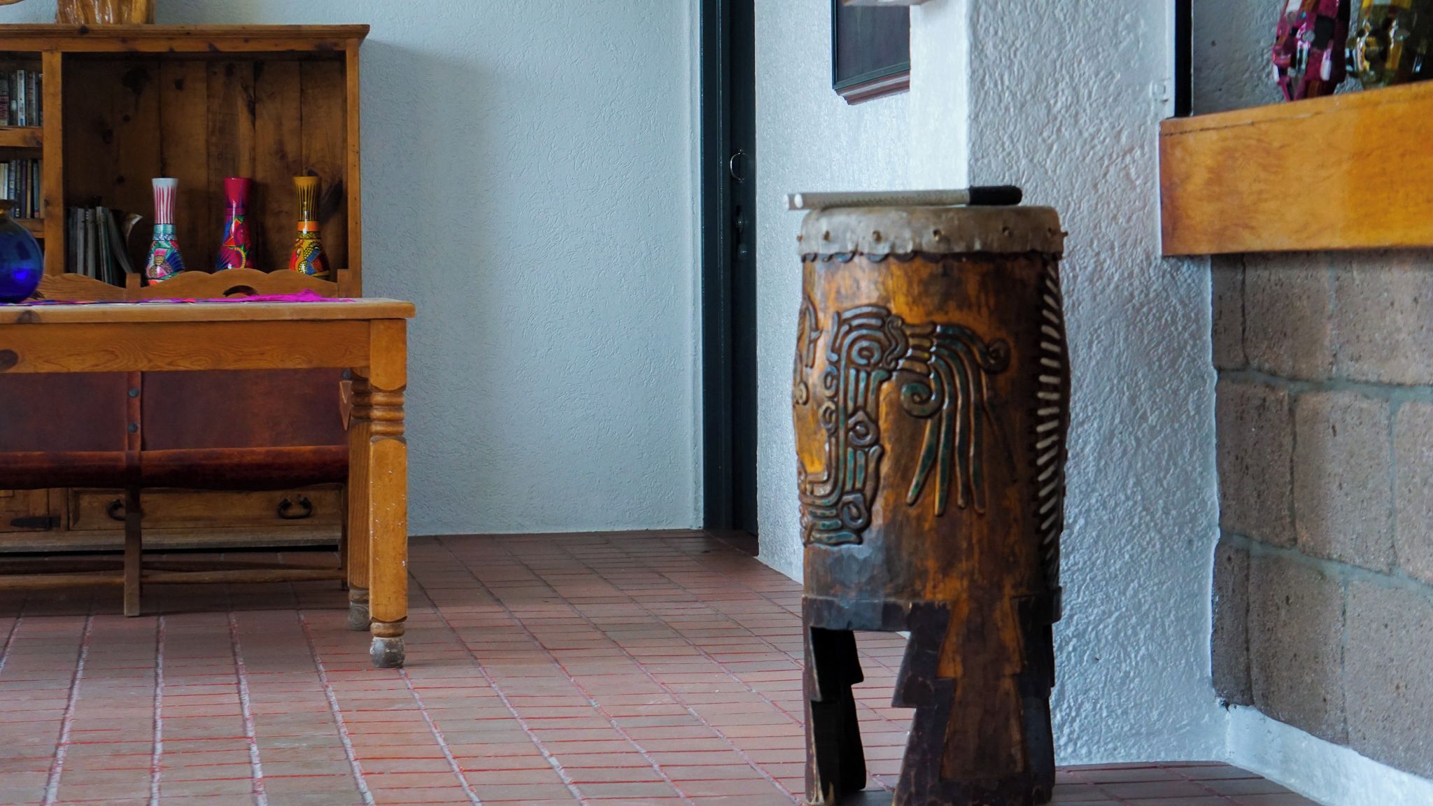 A wooden table with drawers and a decorative vase, a small wooden stool with intricate carvings, and a brick floor.
