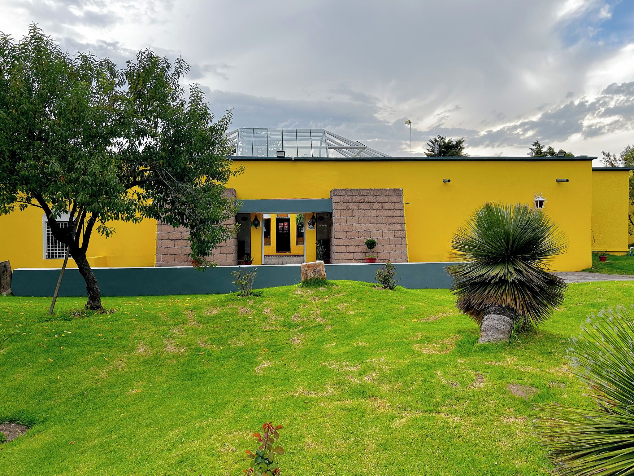 Un edificio amarillo con techo verde, entrada de ladrillo y un pequeño jardín con césped y un árbol.