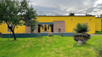 A yellow building with a green roof, a brick entrance, and a small garden with grass and a tree.