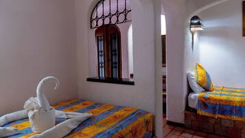 Habitación con dos camas, una con colcha estampada y la otra lisa. Se ve una ventana con celosía metálica y una lámpara de pared.