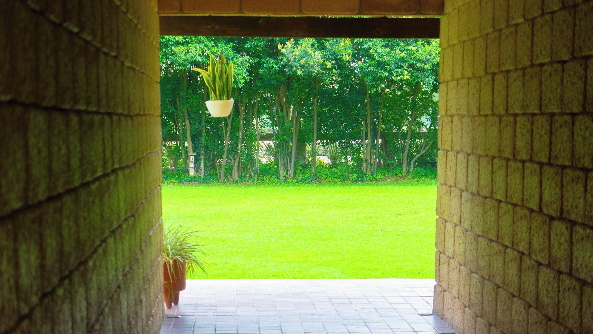 A view through a brick archway into a grassy area with trees and a hanging plant.