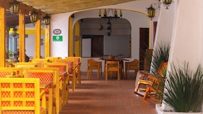 Un espace extérieur aménagé avec des chaises et des tables en bois, un sol carrelé et un mur blanc orné d’une enseigne de bar.