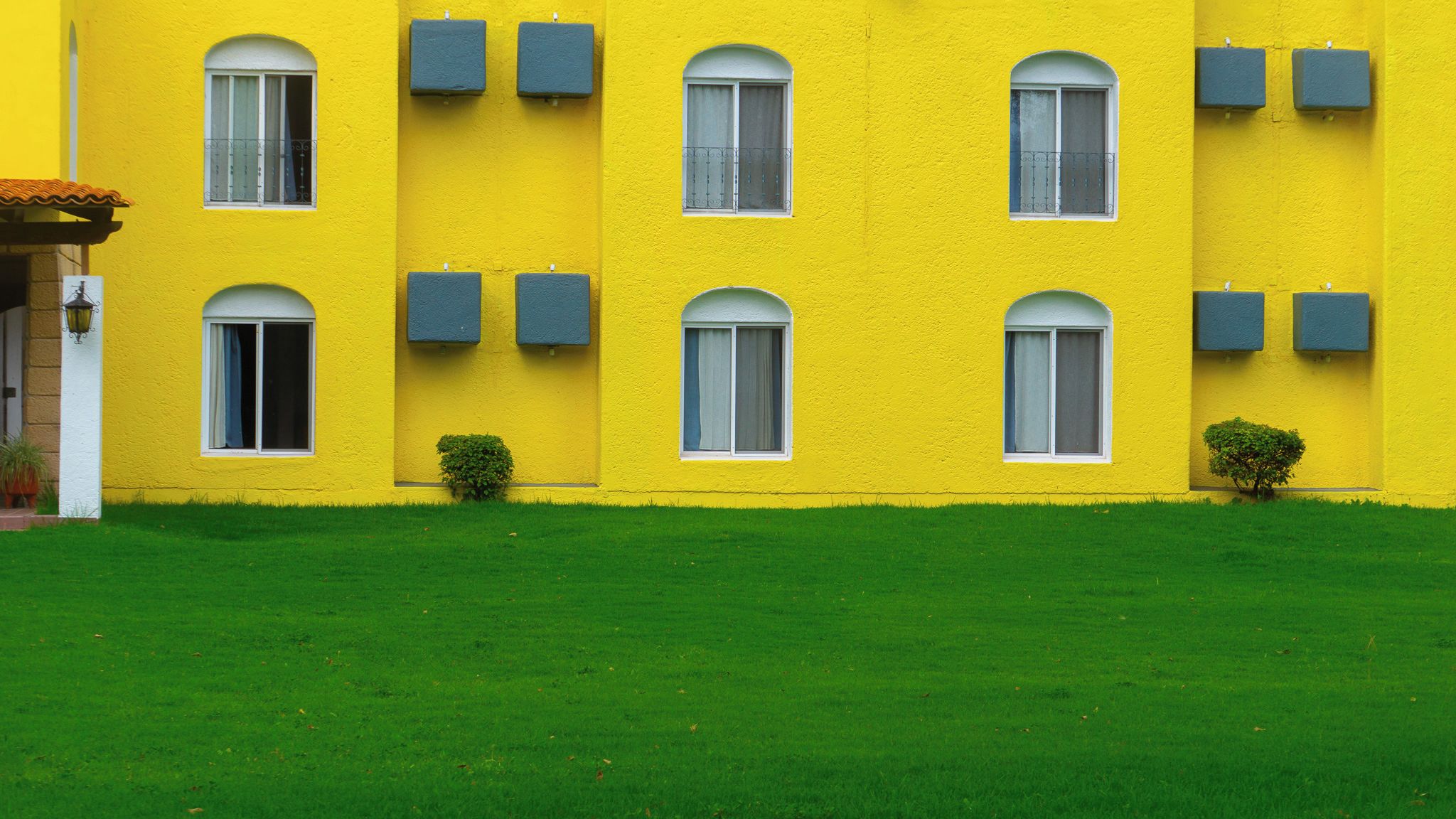 Un edificio amarillo con múltiples ventanas y contraventanas, un césped bien cuidado y un pequeño arbusto.