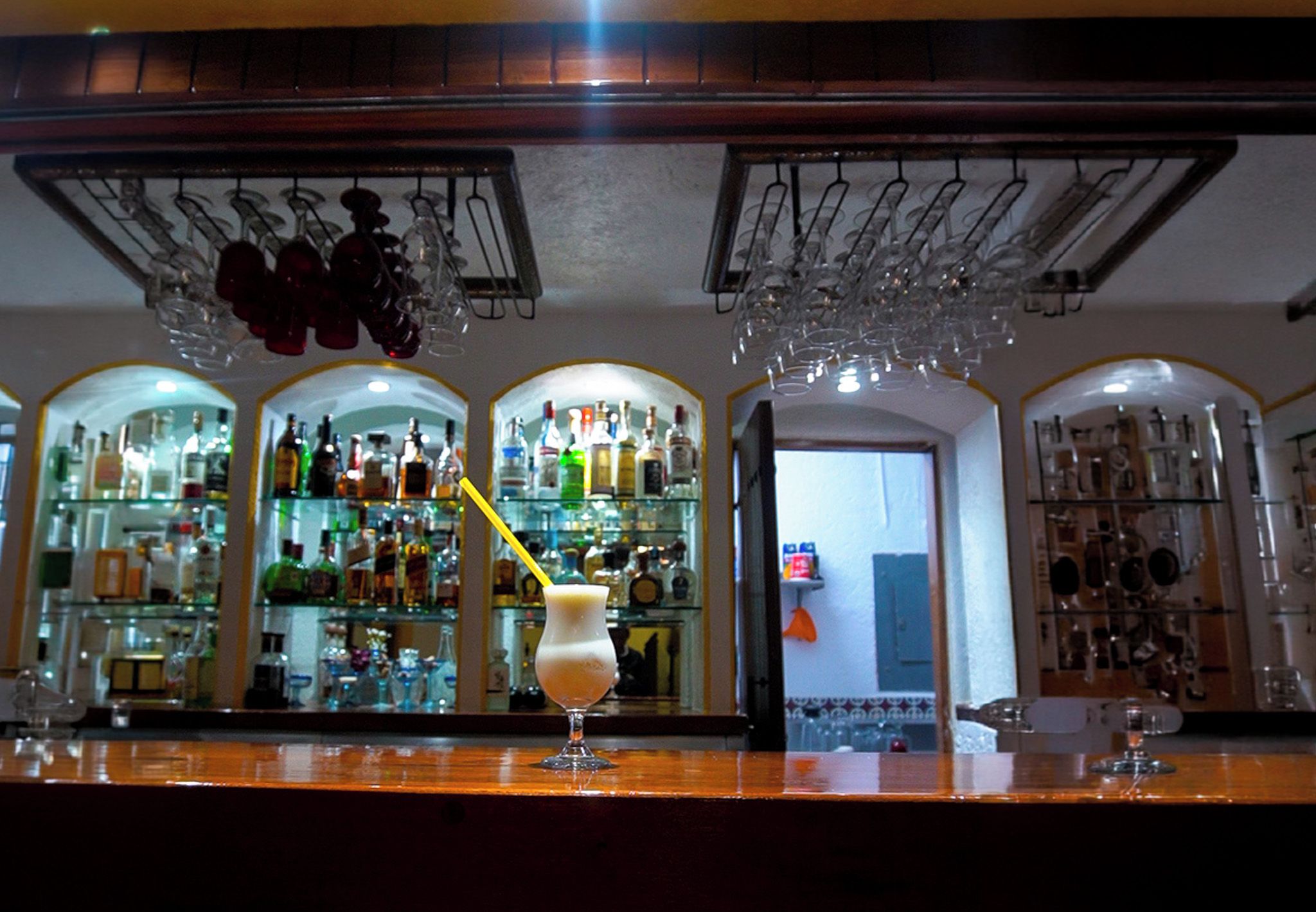 Una zona de bar con estantes llenos de botellas de licor, un vaso con pajita amarilla y un espejo que refleja la barra.