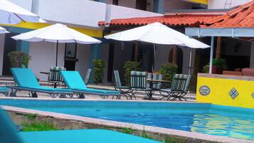 Un área de piscina con sillones de playa y sombrillas, un borde de piscina con azulejos y un edificio con pared amarilla y tejado de azulejos.