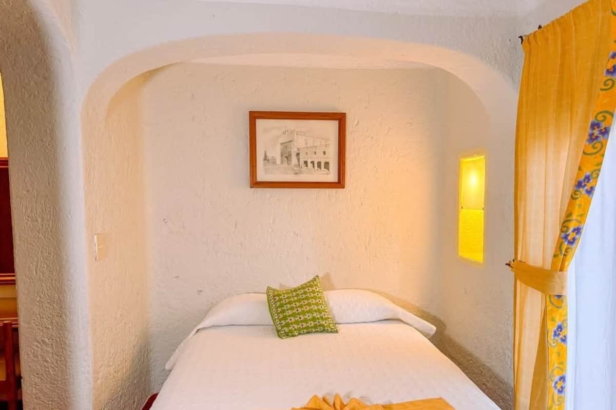 A hotel room with a bed, a framed picture on the wall, and a yellow wall sconce.