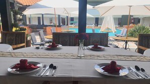 Comedor con mesas dispuestas para cuatro, cada una con un jarrón de flores rojas, cubiertos y servilletas dobladas. Se ven sombrillas y sillones de descanso al fondo, y una piscina es visible.
