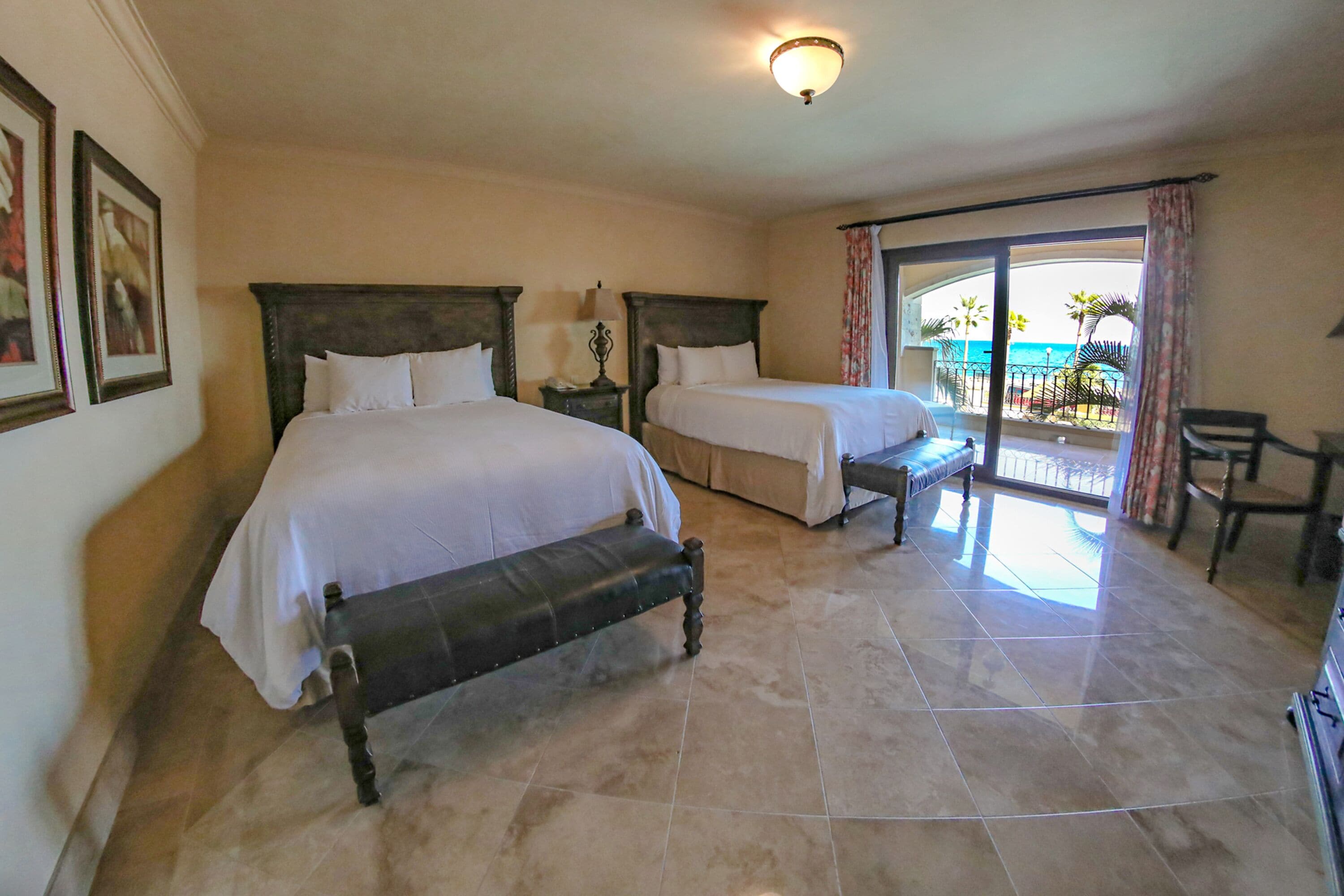A hotel room with two beds, a bench, a desk, and a balcony with a view of the sea.