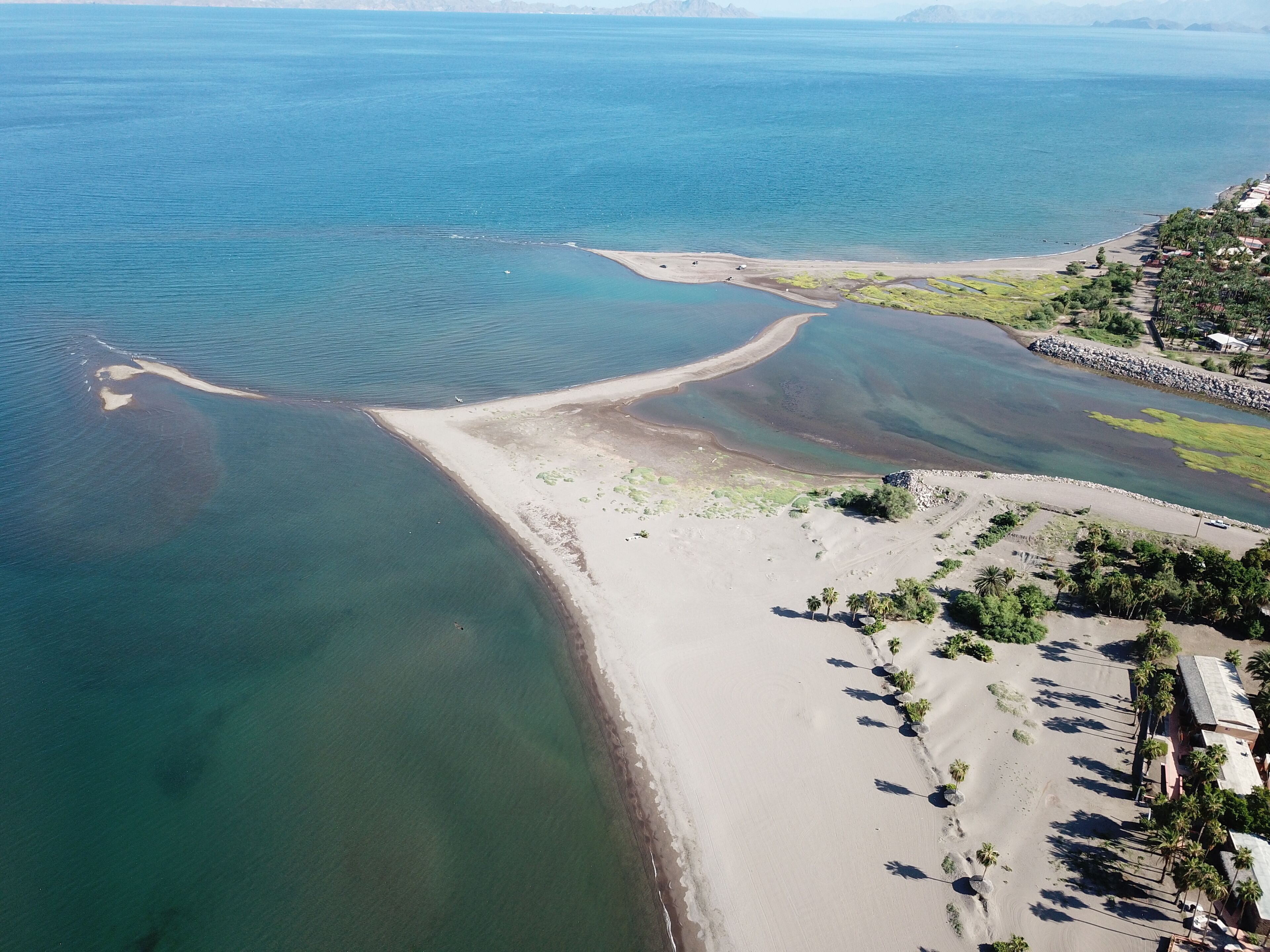 An aerial view of a coastal area with a sandy beach, clear water, and a small peninsula with vegetation.