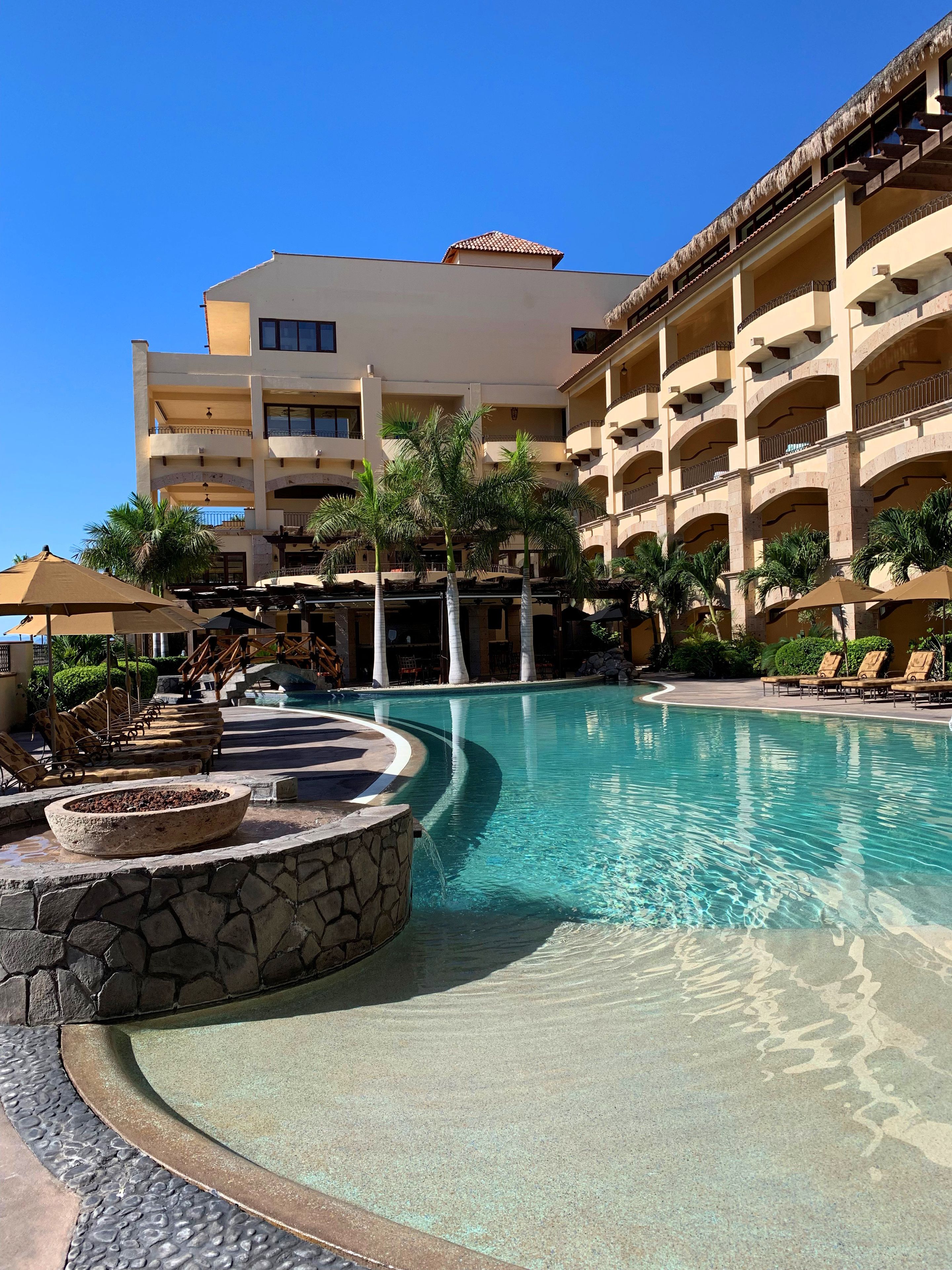 A resort pool area with a clear blue pool, lounge chairs, and a stone fire pit.