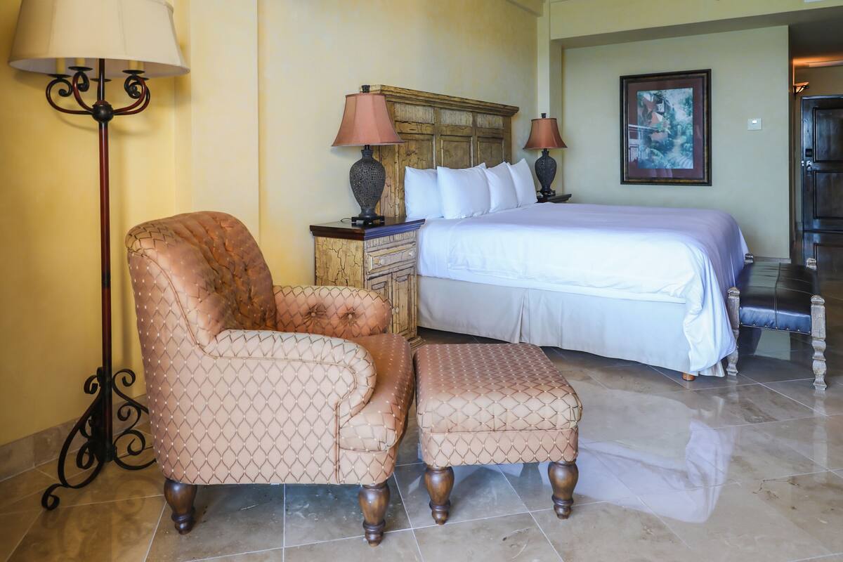 A bedroom with a bed, a chair, an ottoman, a dresser, lamps, and a painting on the wall.