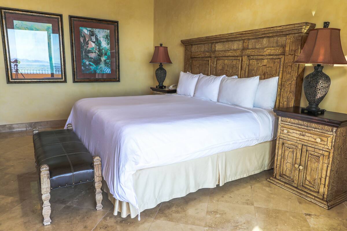 A large bed with a wooden headboard, a bench, two framed pictures, and a nightstand with a lamp.