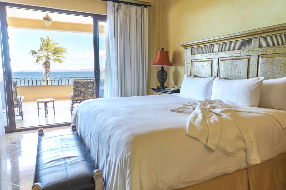 A hotel room with a large bed, a balcony with a view of the sea, a palm tree, and a chair.