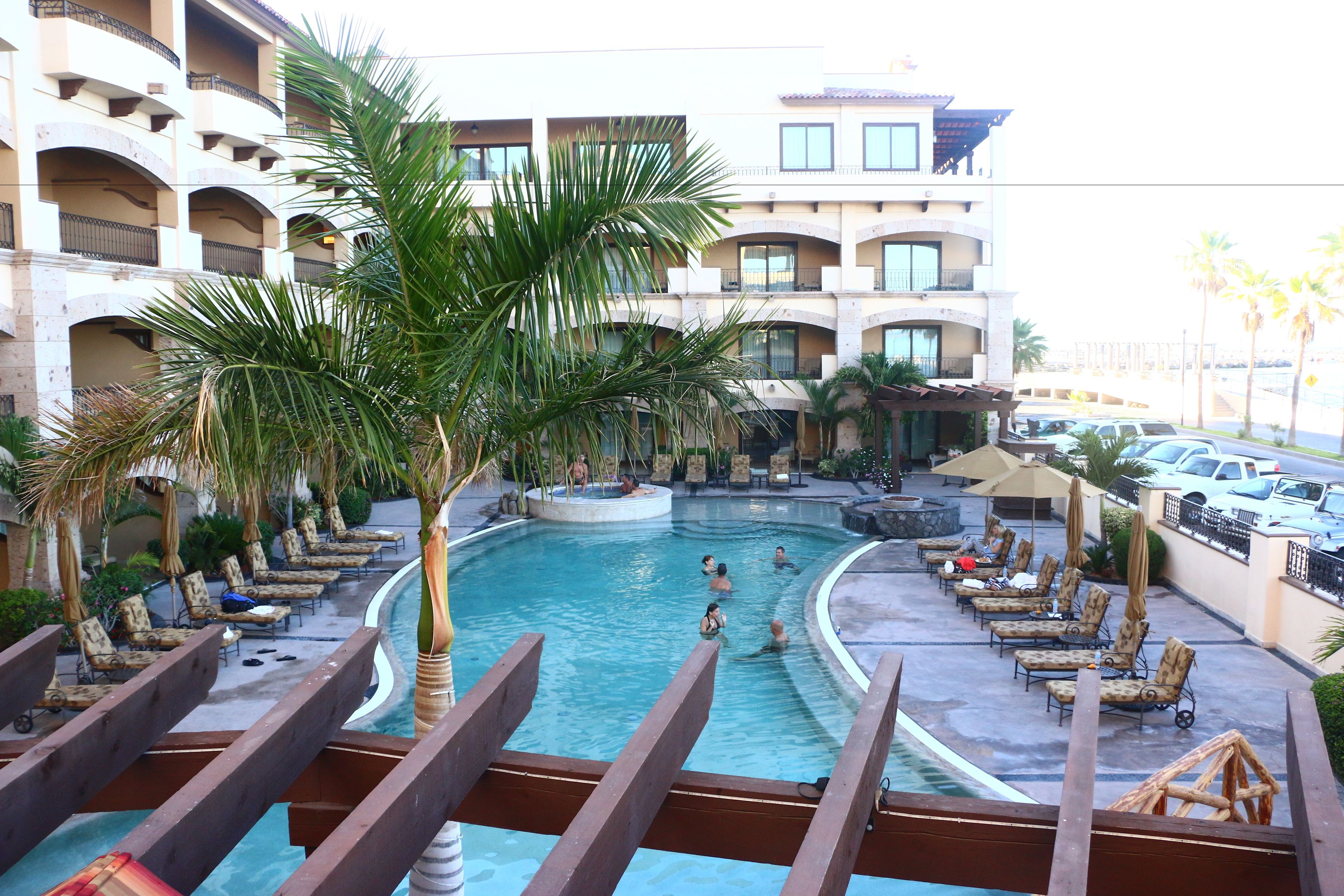 Zona de piscina con sillones de descanso y palmeras, rodeada por un edificio de varios pisos con balcones.