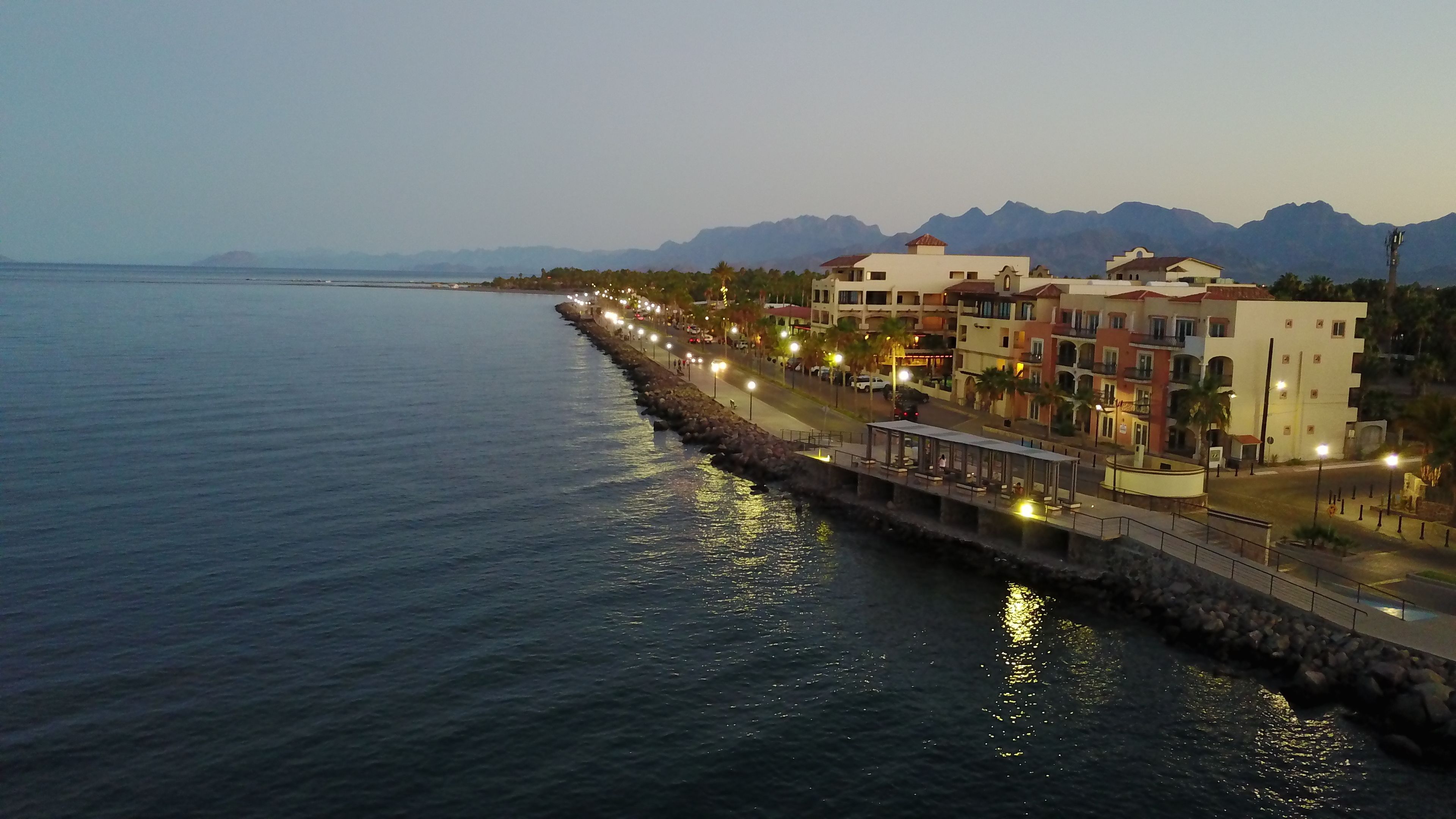 A coastal hotel with a long waterfront promenade, a rocky breakwater, and mountain views in the background.