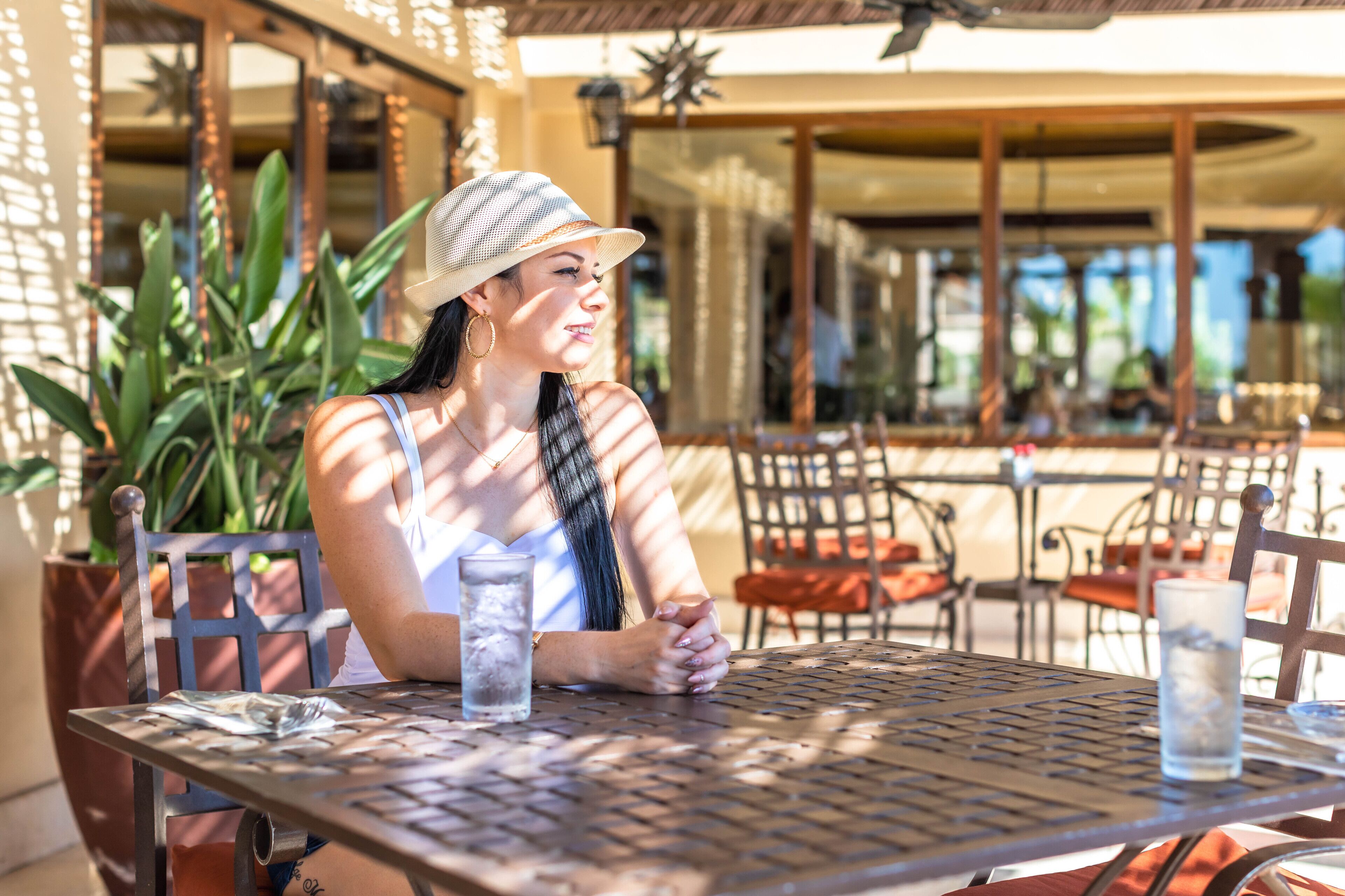 A woman sitting at an outdoor table with a drink, surrounded by chairs and potted plants.