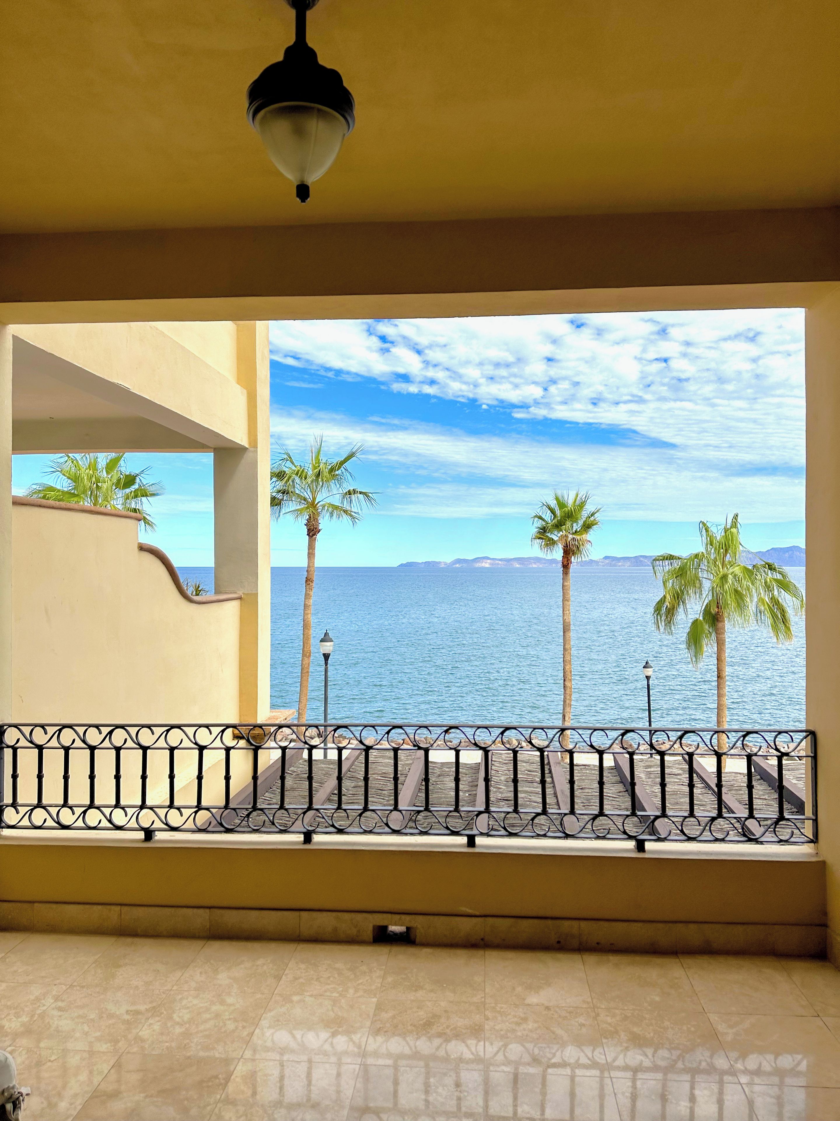 A balcony with a view of the sea, palm trees, and a tiled floor.
