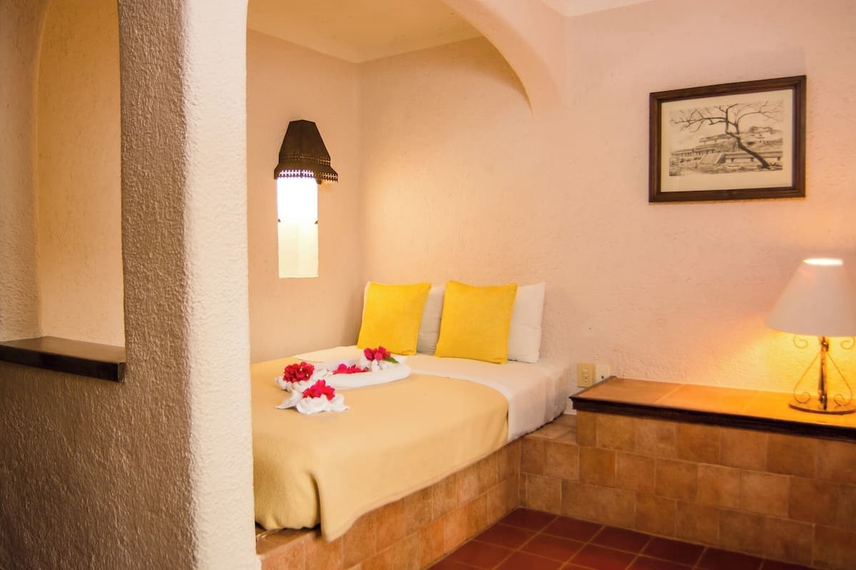 A hotel room with a bed, a nightstand, a lamp, a framed picture on the wall, and a window with a lamp above it.