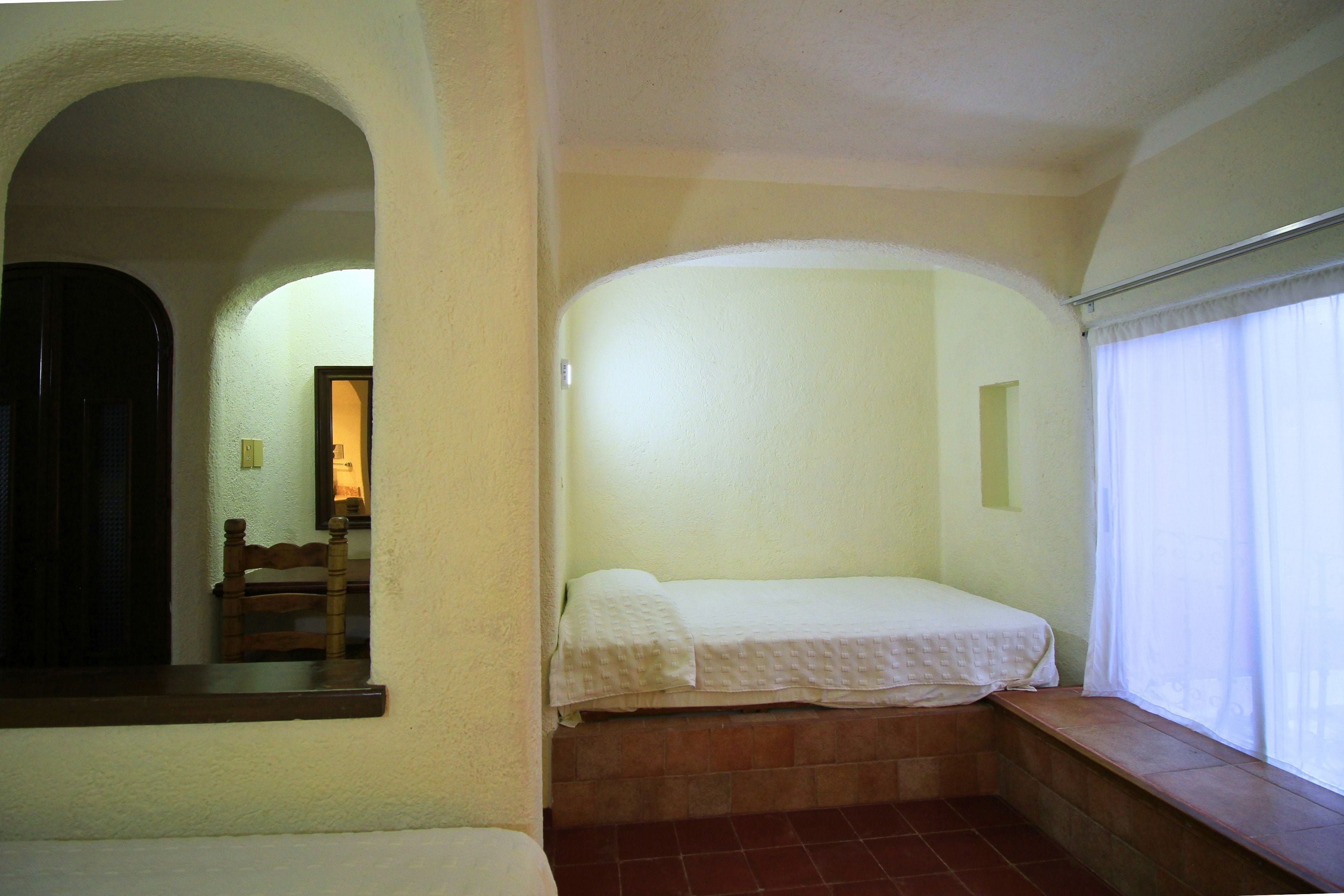 Una habitación pequeña con una cama, un ventanal con cortinas y un banco debajo de la ventana.