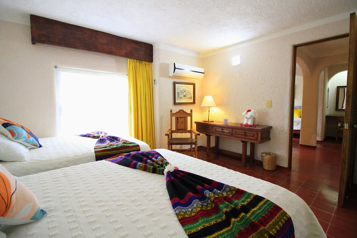 A hotel room with two beds, a desk, a chair, a lamp, and a door leading to another room.