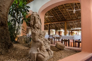 Una escultura de piedra en un entorno tropical con techo de paja y mesas dispuestas para comer.