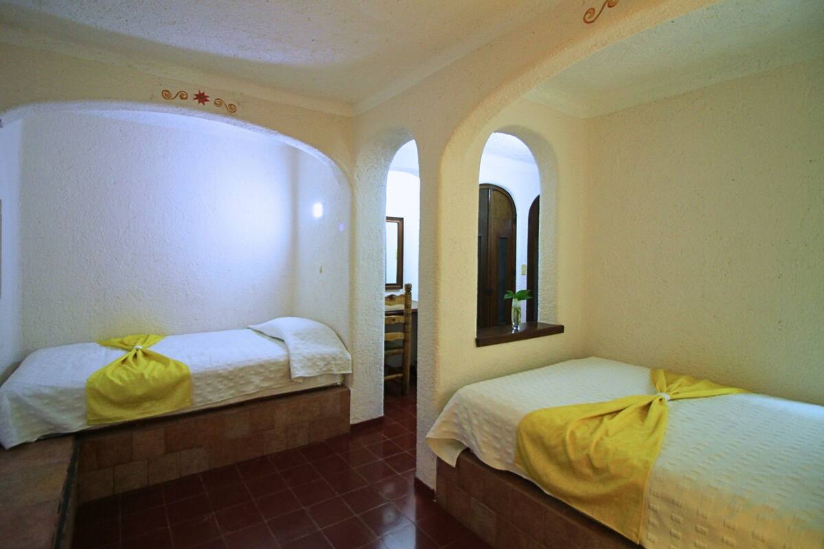 A room with two beds, each with a yellow towel, white bedding, and a mirror above the right bed. The room has a tiled floor and white walls with decorative moldings.