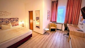 A hotel room with a bed, a small table, a chair, a radiator, and a window with curtains.
