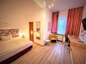A hotel room with a bed, a small table, a chair, a radiator, and a window with curtains. - Arena Hotel (Gelsenkirchen)