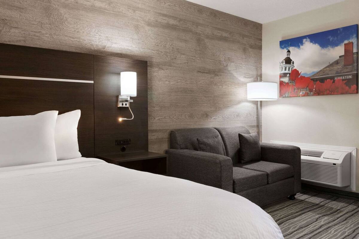 Una habitación de hotel con una cama grande, un sillón gris, una mesita de noche con una lámpara y un cuadro de un edificio.