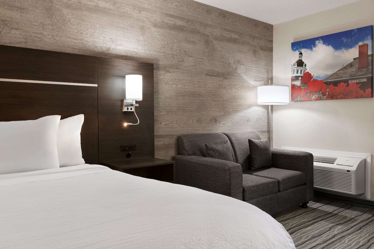 Una habitación de hotel con una cama grande, un sillón gris, una mesita de noche con lámpara y un aire acondicionado instalado en la pared.