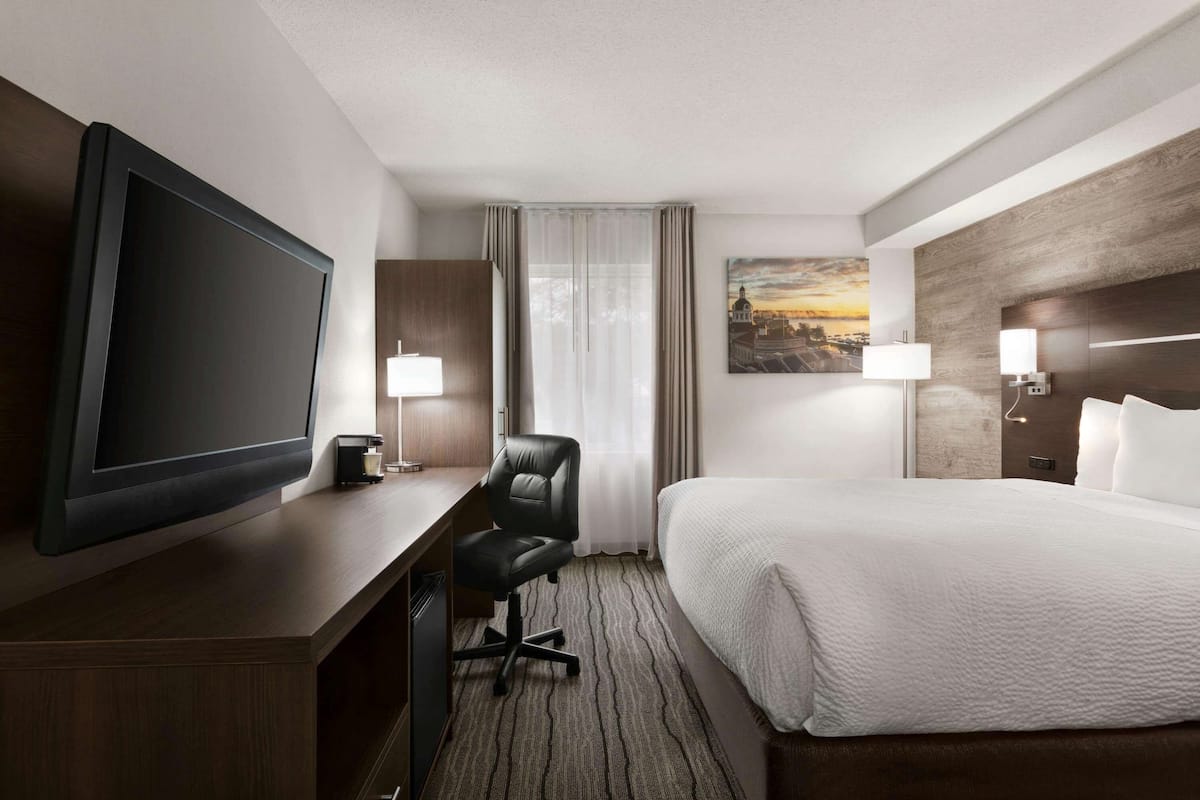 Habitación de hotel con una televisión de pantalla plana grande, un escritorio con silla, una cama con ropa de cama blanca, una ventana con cortinas y un cuadro en la pared.