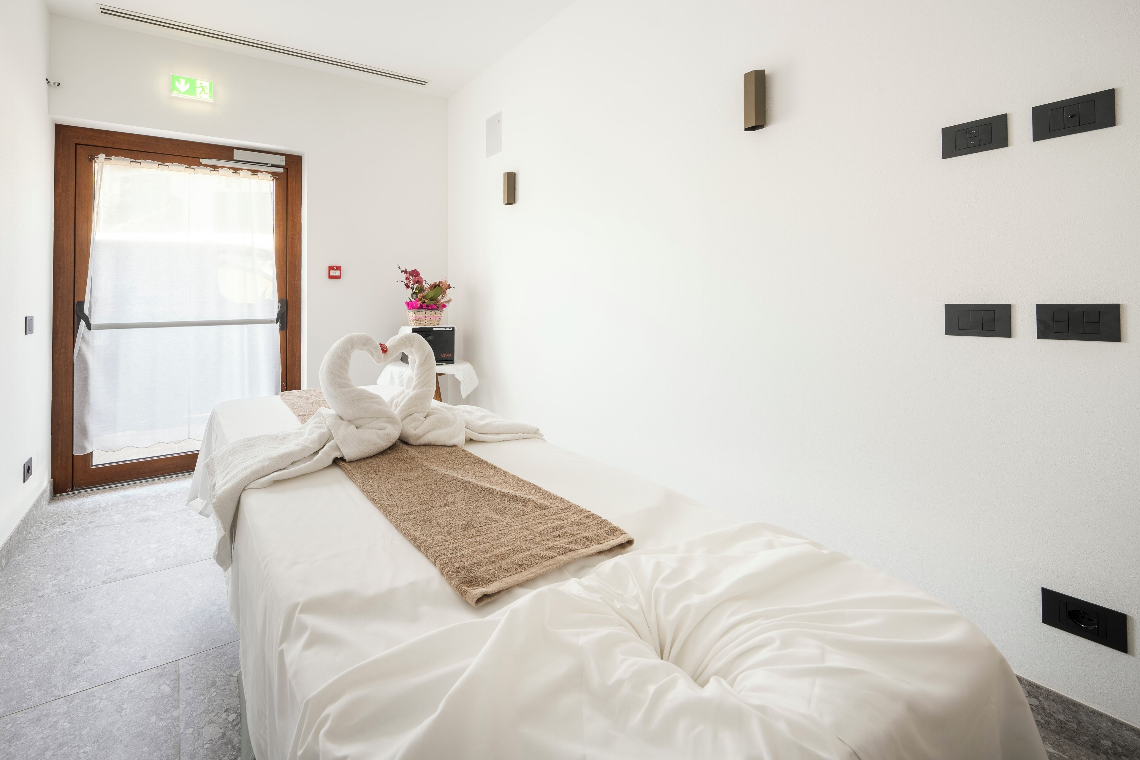 Habitación de hotel con cama, puerta de madera, ventana con cortinas, luz de pared y pequeña mesa con un florero.