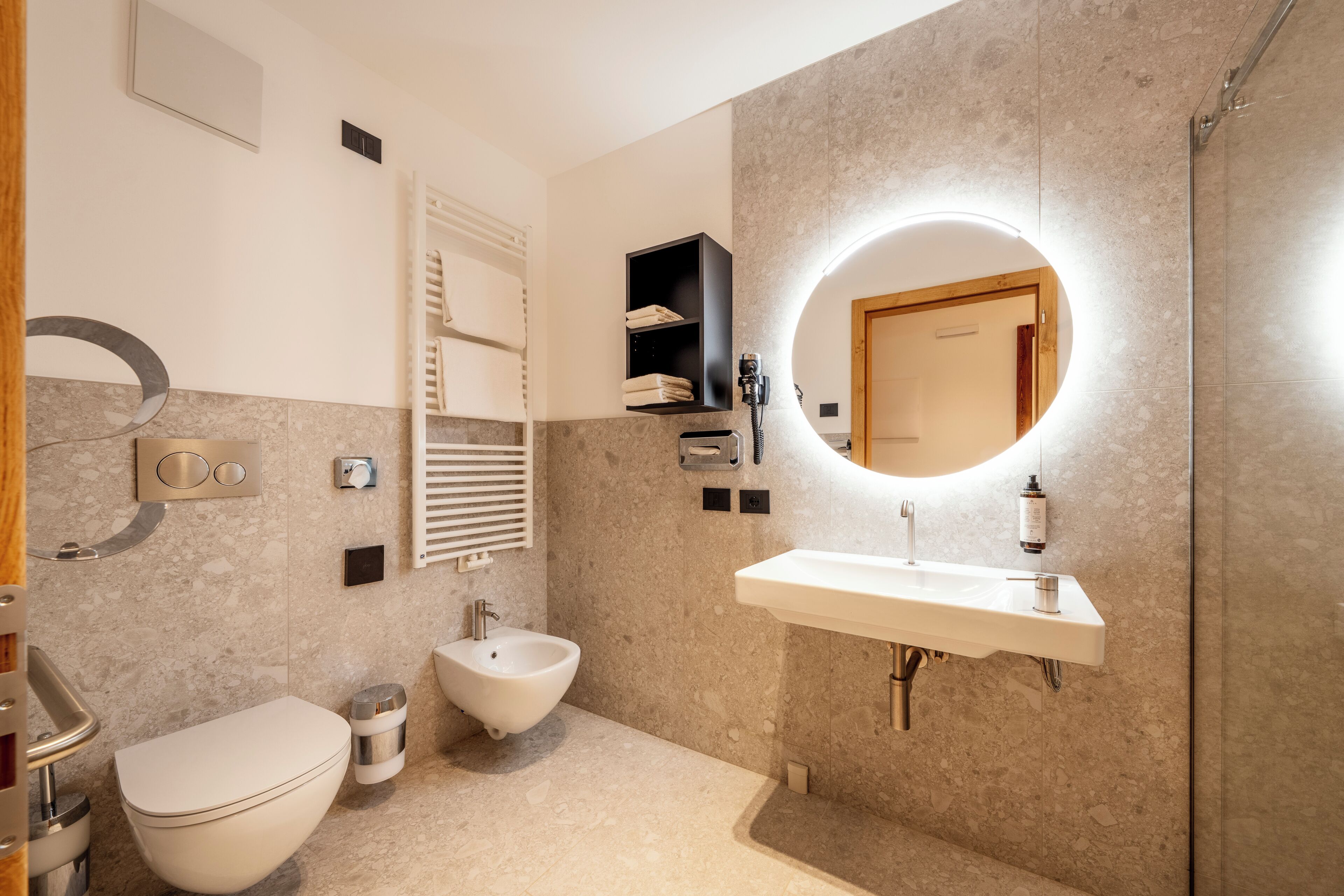 A modern bathroom with a bidet, toilet, sink, and a mirror with a circular light.