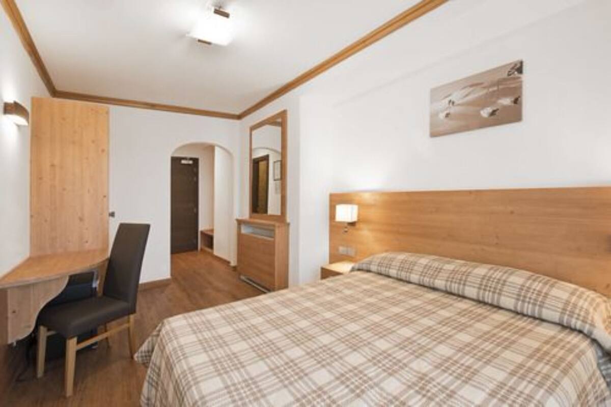 Une chambre à coucher avec une tête de lit en bois, un lit recouvert d’une couverture à carreaux, un bureau avec une chaise, un miroir et un luminaire fixé au mur.